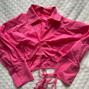 Rosa croppad skjorta med knyt - Säljer en snygg rosa croppad skjorta med knytning framtill. Skjortan har långa ärmar och en klassisk krage. Perfekt för en trendig look!