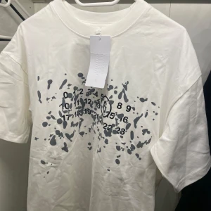 Maison Margiela T-shirt - Säljer en vit t-shirt med ett unikt tryck av siffror och abstrakta former i grått. T-shirten har en rund hals och korta ärmar. Perfekt för en stilren och modern look. Maison margiela t shirt. Nyskick, aldirg använd. Skriv till mig för mer frågor eller bilder🤩💫