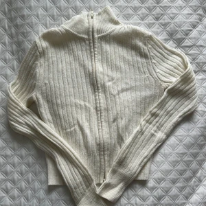 Vit ribbad kofta med dragkedja - Säljer en stilren vit ribbad kofta med dragkedja. Perfekt för kyligare dagar med sin höga krage och långa ärmar. Enkelt att matcha med olika outfits.