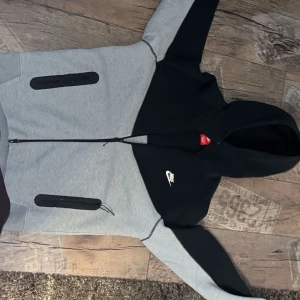 Grå och svart hoodie från Nike - Säljer en snygg grå och svart hoodie från Nike med dragkedja och huva. Jackan har två praktiska fickor med dragkedja på framsidan och Nike-logga på bröstet. Perfekt för en sportig look! Pris går att diskuteras!!