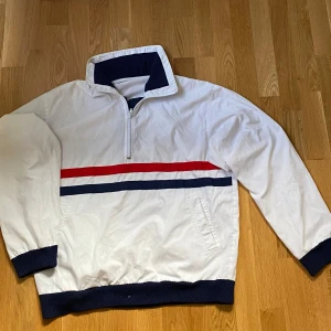 Retro vindjacka/windbreaker - Säljer en vit vindjacka med dragkedja, krage och fickor. Jackan har en röd och en blå rand över bröstet samt blå ribbade muddar vid ärmslut och nederkant. 