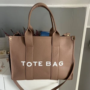 Brun axelväska TOTE BAG liknande 35*28*15 - Snygg brun axelväska med texten 'TOTE BAG' i vitt. Väskan har både handtag och en avtagbar axelrem för flexibilitet. Tillverkad i syntetmaterial med en praktisk dragkedja upptill. Perfekt för att bära med sig allt man behöver under dagen.