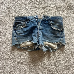 Blå jeansshorts från Levi's - Säljer ett par coola blå jeansshorts från Levi's med fransiga kanter och slitna detaljer. Shortsen har en klassisk femficksdesign och knappgylf. Perfekta för en avslappnad sommarlook!