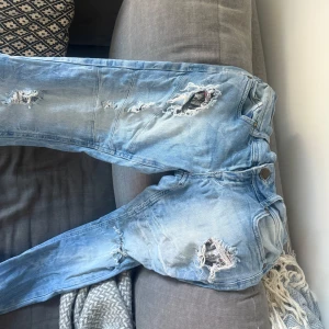 Blå slitna jeans från Fishbone - Säljer ett par blå jeans från Fishbone med slitna detaljer och hål för en trendig look. Jeansen har en knappgylf och klassisk femficksdesign. Perfekta för en trendig stockholmsstil 