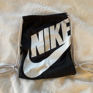 Svart gympåse från Nike - Säljer en svart gympåse från Nike med stort vitt logotryck. Påsen har vita dragsnören och en praktisk stor ficka med dragkedjan på sidan för extra förvaring. Perfekt för träning eller som en lätt ryggsäck för vardagsbruk.