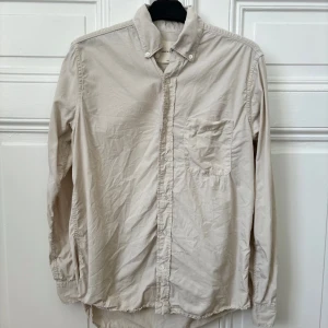 Beige tencel skjorta A Day’s March - Säljer en stilren beige långärmad skjorta med knappar och en bröstficka. Perfekt för en avslappnad men ändå uppklädd look. Skjortan har en klassisk krage och är tillverkad i ett bekvämt material.