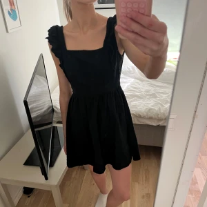 Svart klänning med volangdetaljer - Säljer en söt svart klänning med volangdetaljer vid axlarna. Klänningen har en fyrkantig urringning och är ärmlös, och öppen rygg.