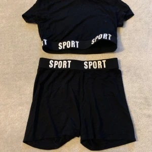 Svart croppad sporttopp med ett par matchande shorts  - Snygg svart croppad sporttopp med elastiskt band där det står 'SPORT' i vitt. Perfekt för träning eller en avslappnad dag. Matcha med de tillhörande shortsen för en komplett look.