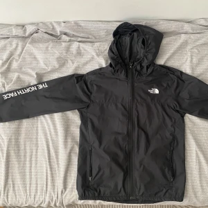 Svart vindjacka från The North Face - Säljer en svart vindjacka från The North Face med huva och dragkedja framtill. Jackan har långa ärmar med logotypen tryckt på ena ärmen och bröstet. Perfekt för blåsiga dagar.(priset kan diskuteras)