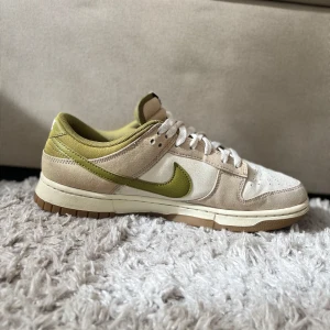 Nike Dunk  - Snygga Nike Dunk sneakers i en stilren kombination av beige och olivgrön. Skorna har en klassisk design med snörning och en bekväm sula. Perfekta för en avslappnad stil.