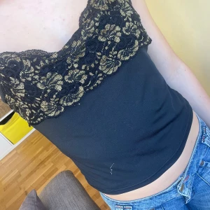 Svart linne med spetsdetaljer - Snyggt svart linne med vackra spetsdetaljer i guld på bröstet. Linnet har smala axelband och en tight passform. Perfekt för en kväll ute eller en speciell tillställning.