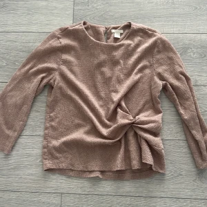 Beige topp med knytdetalj - Säljer en stilren beige topp med en unik knytdetalj framtill. Toppen har trekvartsärmar och en rund halsringning. Perfekt för en chic och avslappnad look.