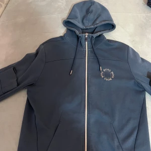 Hugo boss tröja  - Snygg mörkblå huvtröja från Hugo Boss med dragkedja och justerbar huva. Tröjan har ett stilrent tryck med BOSS-loggan på bröstet och ribbade muddar vid ärmslut.