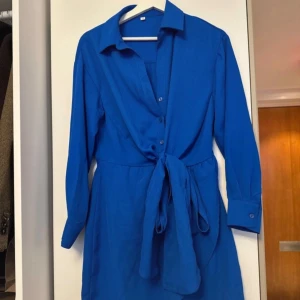 Blå klänning med knytband - Snygg blå klänning med knytband i midjan och långa ärmar. Den har en klassisk krage och knappar framtill. Perfekt för en stilren look.