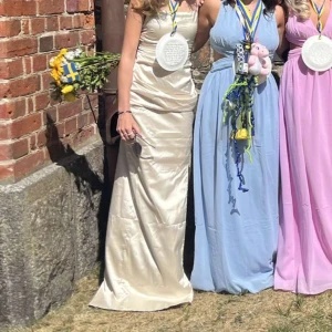Champagnefärgad långklänning - Elegant champagnefärgad långklänning med en slät och glansig yta. Klänningen har en figurnära passform och är ärmlös, vilket ger en stilren och sofistikerad look.