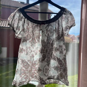 Mönstrad blus med spetsdetalj - Charmig blus med ett detaljerat paisleymönster i dämpade färger. Blusen har korta ärmar och en vacker spetskant vid halsringningen, prydd med en liten rosett. Sjukt söt!