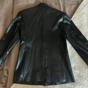 Svart skinnkavaj - Snygg svart kavaj i skinnimitation med knappar framtill. Den har en klassisk design med långa ärmar och en stilren look. Perfekt för att ge en edgy touch till din outfit.✨ stor i strl men passar till XS ( kanske S) 