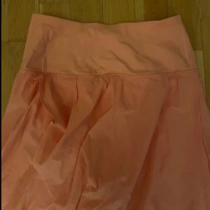 Persikofärgag/rosa kjol från Halara - Snygg persikofärgad/rosa kjol från Halara med en mjuk och flödande design. Kjolen har en hög midja och är perfekt för en avslappnad stil. Materialet är lätt och bekvämt, vilket gör den idealisk för varma dagar.