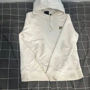 Vit hoodie från Lyle & Scott - Säljer en stilren vit hoodie från Lyle & Scott med deras ikoniska logga på bröstet. Tröjan har en klassisk känguruficka och ribbade muddar vid ärmslut och nederkant. Perfekt för en avslappnad look.