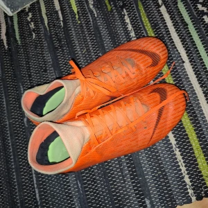 Orangea Nike fotbollsskor med dobbar - Säljer ett par orangea Nike fotbollsskor med svarta detaljer och snörning. Skorna har dobbar för spel på gräs och en smal, sportig design. Perfekta för dig som vill sticka ut på planen med stilrena och snabba skor.