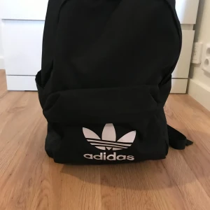 Svart Adidas ryggsäck Primegreen - Svart ryggsäck från Adidas med stort vitt Trefoil-logga framtill. Tillverkad i slitstark polyester och märkt med Primegreen, vilket innebär återvunnet material. Rymligt huvudfack, framficka med dragkedja och justerbara axelremmar. Perfekt för skola eller träning. Aldrig använd 
