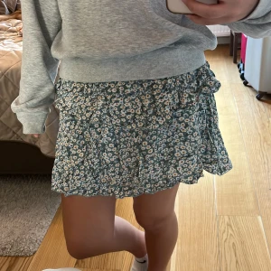 Blommig volangkjol i grönt och vitt - Supersöt kort kjol med blommigt mönster i vitt, gult och blått på grön botten. Kjolen har volangdetaljer och resår i midjan för en bekväm passform. Perfekt för sommaren🥰