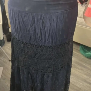 Mörkblå långkjol med spetsdetaljer - Snygg mörkblå långkjol med bred midja och vackra spetsdetaljer i mitten. Kjolen har en lätt rynkad struktur och faller fint ner mot anklarna. Perfekt för dig som gillar boho-stil och vill ha något unikt i garderoben.