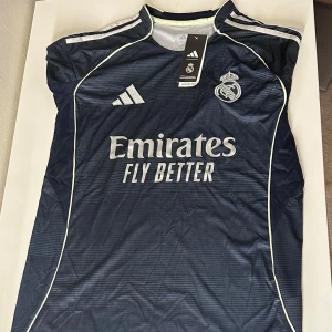 Real Madrid bortatröja Adidas 25/26 - Snygg Real Madrid bortatröja från Adidas i mörkblått med tunna horisontella ränder. Bästa kvaliten finns bara hos oss på BallerDrip, Alla storlekar finns