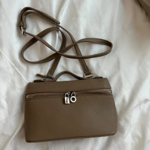 Beige Loro Piana axelväska med dragkedja - Beige Loro Piana axelväska i strukturerat skinn med justerbar rem. Väskan har en praktisk dragkedja framtill med silvriga detaljer och en minimalistisk design som passar till många outfits.