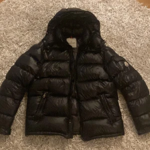 Svart Moncler dunjacka med huva - Säljer en svart, glansig dunjacka från Moncler med huva och dragkedja framtill. Jackan har två snedställda fickor med dragkedja och klassisk Moncler-logga på ärmen. Perfekt för kalla dagar och riktigt snygg streetstil. Inga repor eller skador. Riktig moncler Maya 