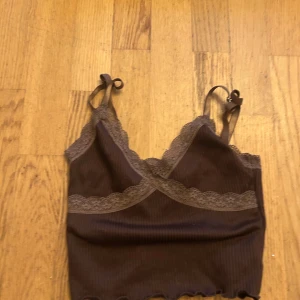 Brun ribbad linnetopp med spets - Supersöt brun ribbad linnetopp med tunna axelband och fina spetsdetaljer längs kanterna. Toppen har små rosetter på axelbanden och en vågig kant nertill som ger en extra touch. Perfekt att styla med jeans eller kjol för en chill look.