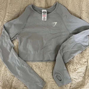 Grå croppad träningströja Gymshark - Snygg grå croppad långärmad träningströja från Gymshark. Tröjan har tumhål i ärmsluten, diskret mönster i tyget och en tight passform. Materialet är en blandning av nylon och elastan för maximal stretch och komfort. superskön. 