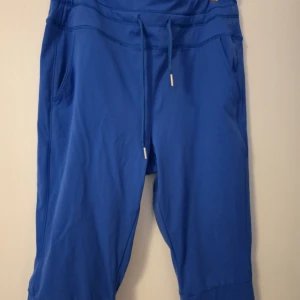 Blå träningsbyxor med snörning - Snygga blå träningsbyxor i trekvartsmodell med dragsko i midjan och fickor på sidorna. Tillverkade i stretchigt material av 77% nylon och 23% elastan, perfekta för träning eller chill. Regular fit och sportig look. Nya, bara provade. Katt finns I hemmet