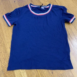 Marinblå ribbad topp från H&M XS - En marinblå ribbad topp från H&M i storlek XS. T-shirten har korta ärmar och sportiga ränder i vitt och rött vid halsringning och ärmslut. Perfekt för dig som gillar en enkel men ändå färgglad stil.