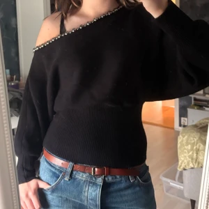Svart offshoulder stickad tröja med pärlor - Svart stickad tröja från ZARA med bred ribbad midja och offshoulder-modell. Snygg detalj med stora silvriga pärlor längs den asymmetriska halsringningen.
