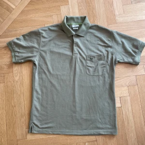Olivgrön pikétröja från Lacoste - Säljer en olivgrön pikétröja från Lacoste med klassisk krage, korta ärmar och bröstficka med den ikoniska krokodilloggan. Tröjan är i mjuk bomull och har en stilren, enkel design som passar perfekt till jeans eller shorts.