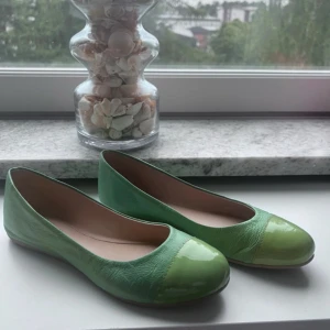 Gröna ballerinaskor från Inkognito - Snygga ballerinaskor i två nyanser av grönt från Inkognito. Skorna har rund tå och en glansig detalj framtill som ger en cool kontrast till det matta skinnet. Perfekta för dig som vill sticka ut med färg på fötterna.