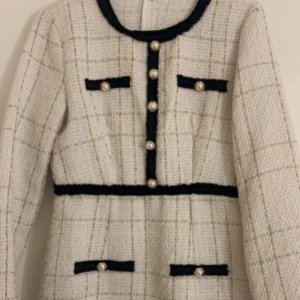 Vit rutig klänning med pärlknappar - Supersnygg vit klänning med rutigt mönster och svarta detaljer. Klänningen har långa ärmar, dekorativa pärlknappar framtill och på fickorna samt markerad midja. Perfekt för dig som gillar en chic och klassisk stil.