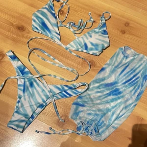 Blåvit bikini med snörning och meshkjol - Säljer en snygg bikini i blått och vitt med tie-dye mönster. Bikinin har trekantsöverdel med långa knytband och matchande trosa. Ingår även en meshkjol med dragsko i samma mönster. Perfekt för stranden eller poolen!