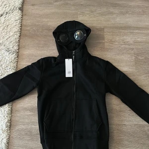 Svart hoodie från C.P. Company - Svart hoodie från C.P. Company med ikoniska glasögon i huvan. Hoodien är bara testad och aldrig använd eftersom den va för liten. Storlek M. Nypris cirka 3000 