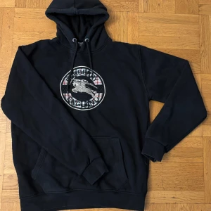 Svart Burberry hoodie med logga - Svart hoodie från Burberry med stor broderad logga och rutig detalj framtill. Klassisk känguruficka, snörning i huvan och mjukt material. Perfekt för dig som gillar streetstyle och vill ha något som sticker ut.