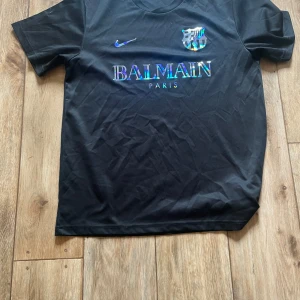 Svart Barcelona x Balmain fotbollströja M Nike  - Snygg svart fotbollströja från Nike med Balmain Paris-tryck och holografiska detaljer på bröstet. Tröjan har korta ärmar och är tillverkad i lätt och ventilerande material (Nike Aeroswift). Perfekt för dig som vill sticka ut på planen eller gatan.