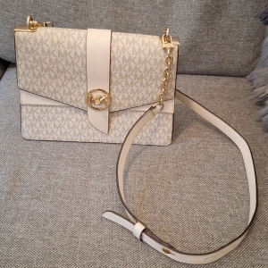 Beige axelväska från Michael Kors - Snygg beige axelväska från Michael Kors med MK-logga över hela väskan. Väskan har guldfärgade detaljer, kedja på axelremmen och ett stilrent spänne framtill. Tillverkad i syntetmaterial med struktur och klassisk rektangulär form.