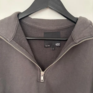 Mörkgrå half zip tröja från 157 - Mörkgrå långärmad tröja från 157 med halv dragkedja och hög krage. Tröjan är i mjuk bomullsmix och har en relaxed passform. Perfekt för lager-på-lager och chill dagar. Enkel och stilren design utan tryck eller mönster.