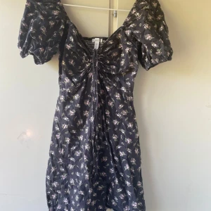 Blommig kortklänning i linneblandning - Supersöt kortklänning med svart botten och små blommor i rosa och grönt. Puffiga korta ärmar, v-ringning och snygg snörning framtill. Tillverkad i linneblandning, perfekt för varma dagar. Sitter fint och ger en romantisk vibe.