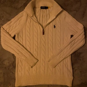 Vit stickad half zip tröja från Ralph Lauren - Säljer en vit stickad tröja från Ralph Lauren med kabelstickat mönster och dragkedja vid halsen. Tröjan har långa ärmar och en broderad logotyp på bröstet. Perfekt för kyligt väder.