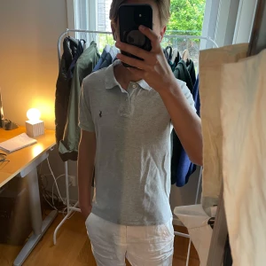 Grå pikétröja från Ralph Lauren - Säljer en stilren grå pikétröja från Ralph Lauren med klassisk krage och knappar framtill. Tröjan har korta ärmar och den ikoniska broderade loggan på bröstet. Perfekt för en clean och avslappnad look.