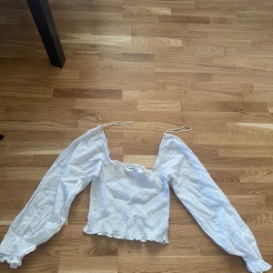 Vit blus med puffärmar från NA-KD - Supersöt vit blus från NA-KD med smockad kropp och långa puffärmar. Toppen har knytband vid axlarna och volangkant nertill. Perfekt för dig som gillar romantisk och trendig stil.