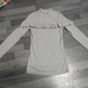 Grå ribbad tröja med volang Chiara Forthi - Supersöt ljusgrå ribbad tröja från Chiara Forthi med volangdetalj över bröstet. Hög hals och långa ärmar ger en stilren vibe. Perfekt att styla med jeans eller kjol för en trendig look.