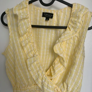 Gul topp från Topshop - Söt gul- och vitrandig topp från Topshop med volangdetaljer längs urringningen. Ärmlös modell i lätt bomullsmaterial, perfekt för varma dagar. Klänningen har en fräsch och somrig vibe med elastisk midja för skön passform.